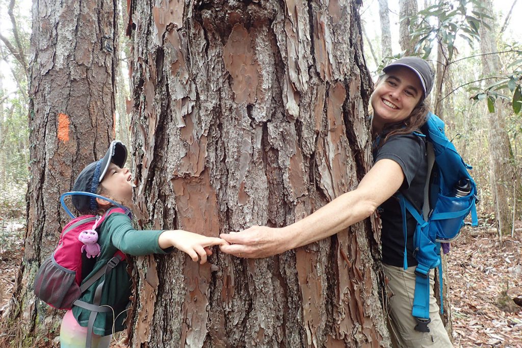 A mother and daughter hug a tree (slash pine) from opposite sides.