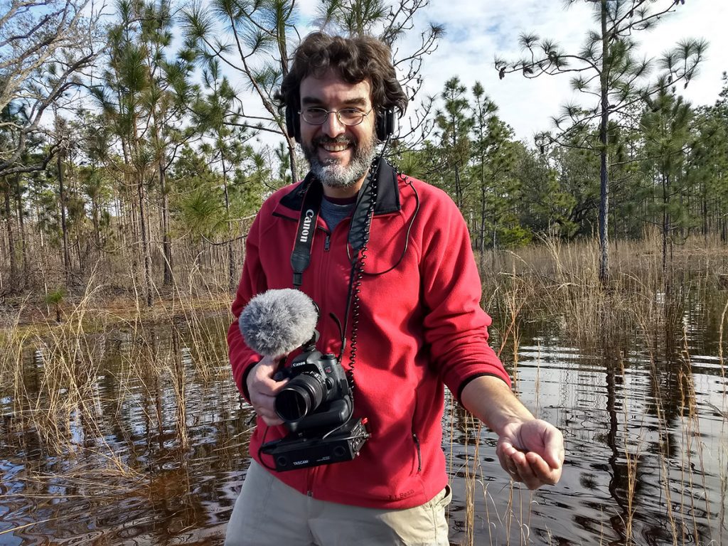 WFSU producer Rob Diaz de Villegas with a camera in one hand, and two striped newts in the other.