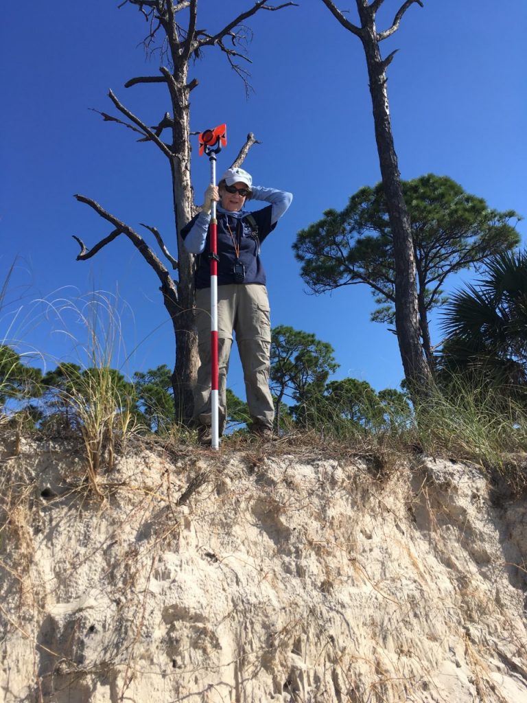 Susan Cerulean stands atop a dune with a monument meant to measure erosion.  The steepness of the escarpment (almost vertical as opposed to a gradual slope) is evidence of shoreline erosion.